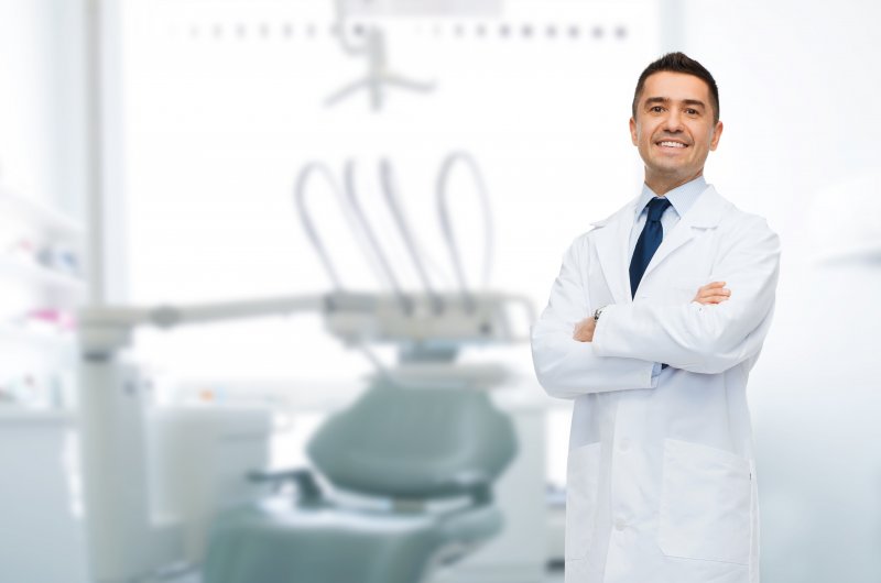 a male dentist standing with his arms crossed and smiling while wearing a lab coat in a treatment room